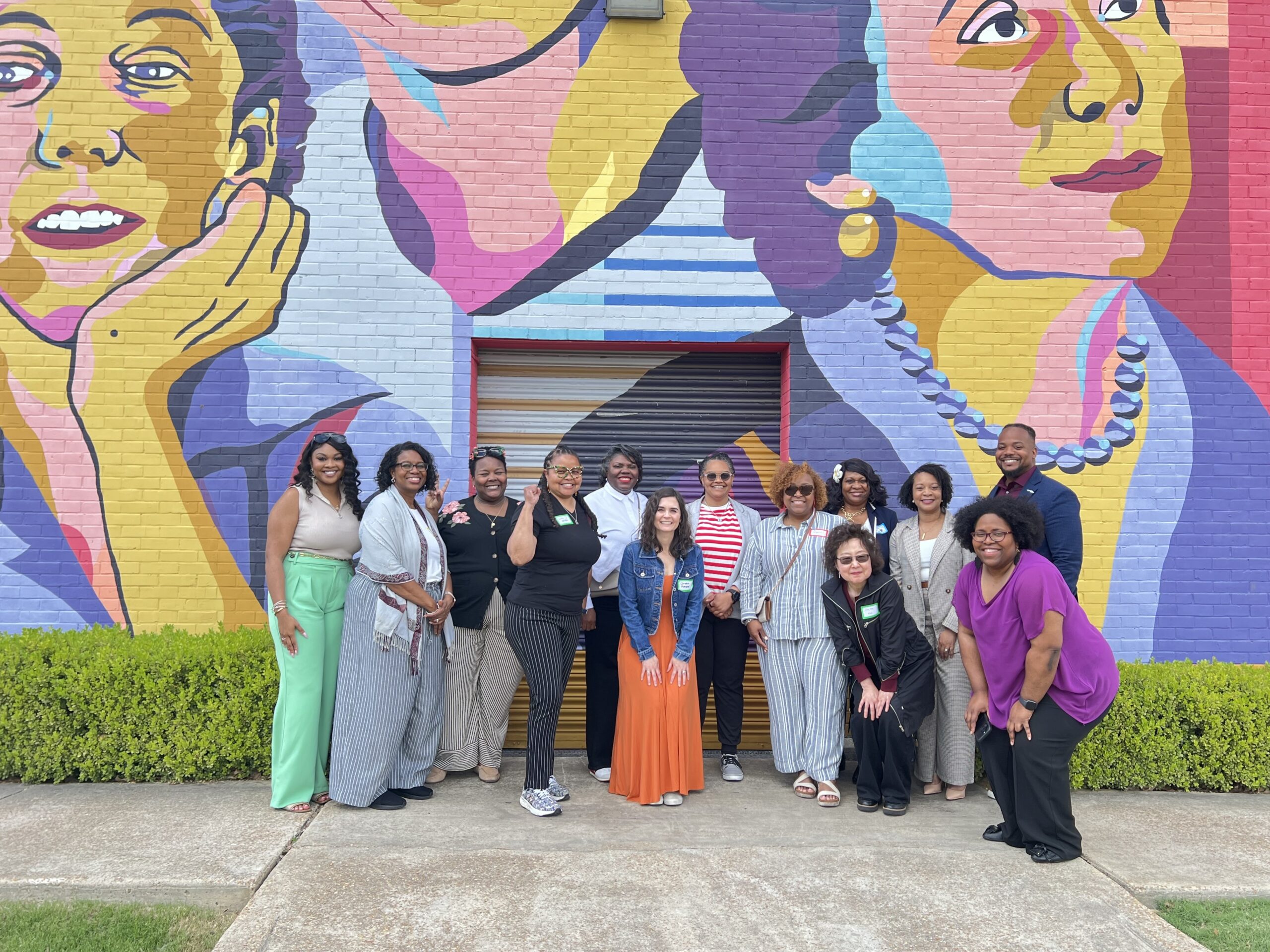 The image shows a large group of thirteen people posing for a photo in front of a vibrant, colorful mural.

  * **Mural details**: The mural on the brick wall behind the group depicts large-scale, stylized portraits of several women in shades of yellow, purple, blue, and red.
  * **Group composition**: The group consists of twelve women and one man, standing and smiling in a single row along a sidewalk.
  * **Attire**: They are dressed in a variety of casual and professional clothing, including dresses, blazers, and patterned trousers.
  * **Setting**: The group is standing outdoors on a light-colored sidewalk in front of a low green hedge at the base of the mural.

